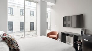 A hotel room with a bed, a desk, a chair, a TV, and a window with a view of buildings.