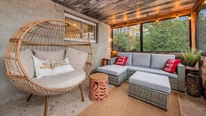 A cozy seating area with a wicker chair, a sofa, and a small round table, all set against a concrete wall with a large window overlooking greenery. - Pôr do Sol  Whitelake Waterfront  Group  Getaway (White Lake)