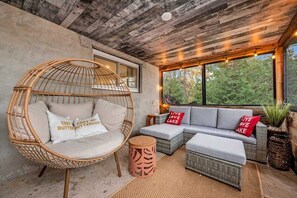 A cozy seating area with a wicker chair, a sofa, and a small round table, all set against a concrete wall with a large window overlooking greenery.