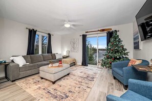 A living room with a grey sofa, a blue armchair, a coffee table, a Christmas tree, and a sliding glass door leading to a balcony.