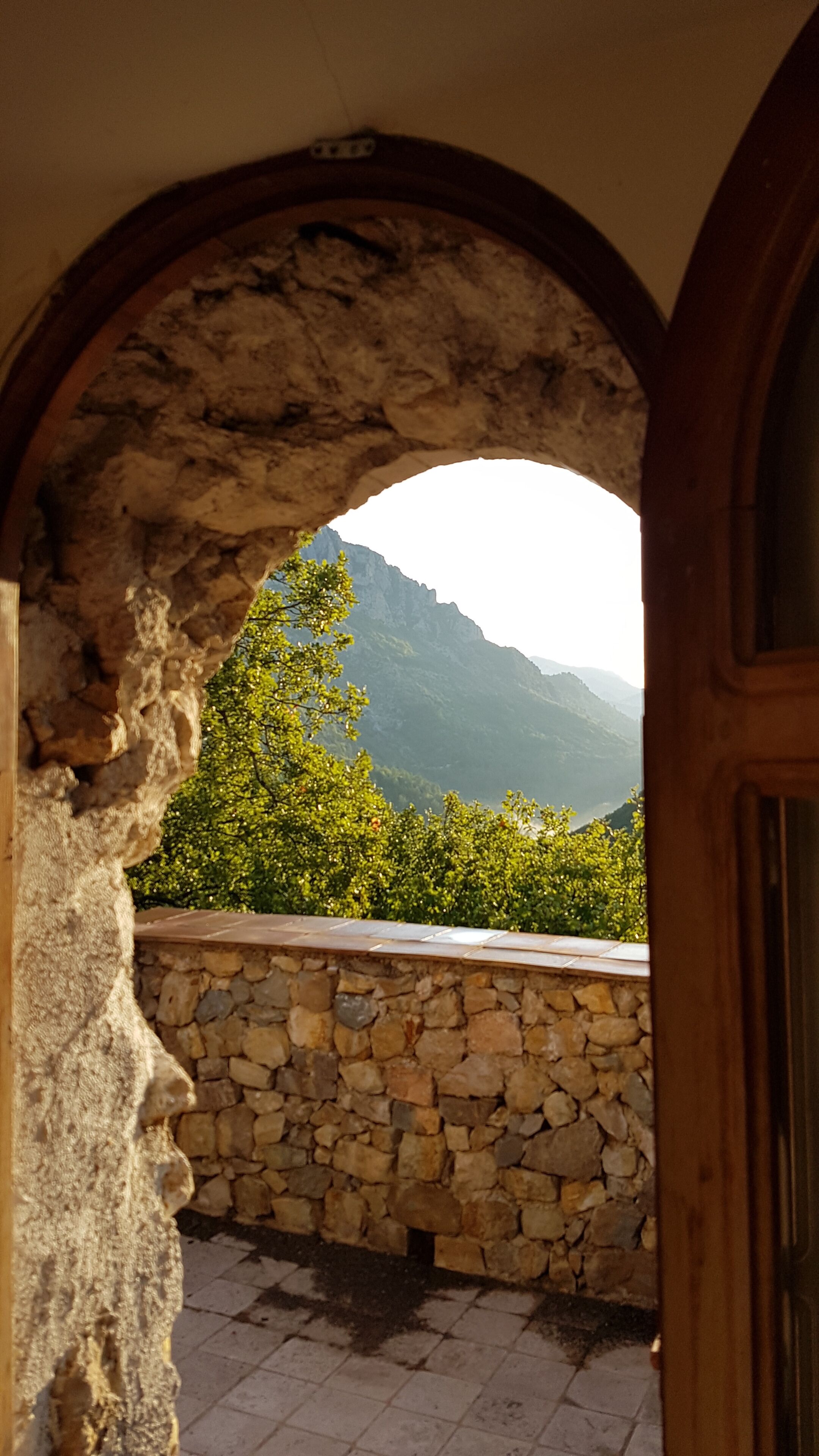 An arched stone doorway leading to a balcony with a stone wall and a view of green trees and mountains.