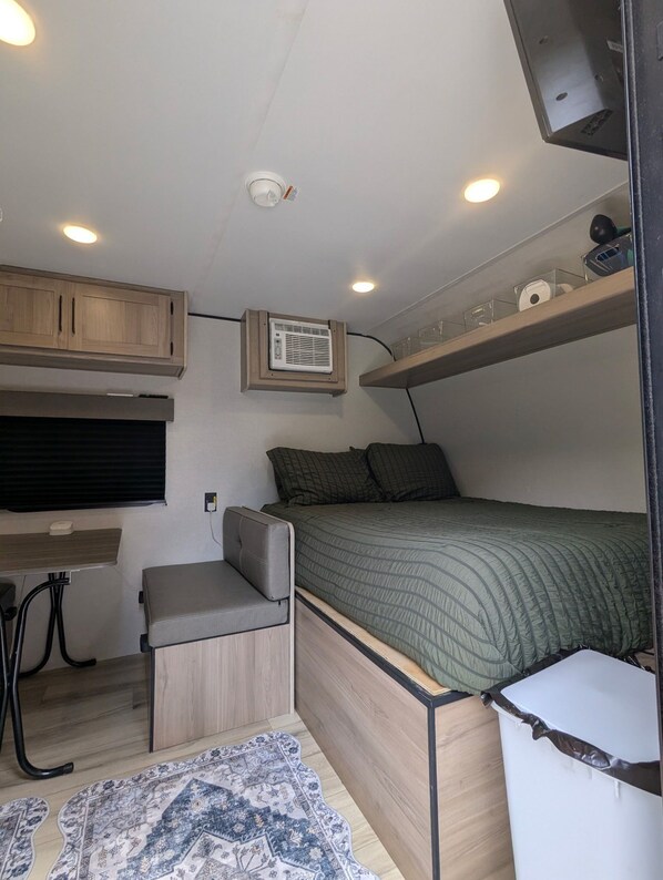 A compact RV interior with a bunk bed, a small desk area, and a ceiling-mounted air conditioning unit.