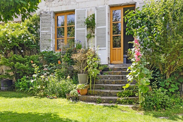 A stone building with a wooden door and shuttered windows, surrounded by lush greenery and potted plants.