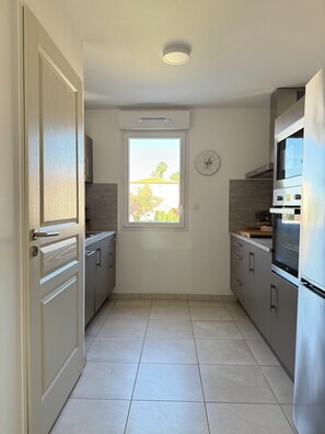 A narrow kitchen with a window, a clock, and modern appliances. - T2 avec terrasse, parking et cellier | La Roselière Le Grau-du-Roi (Le Grau-du-Roi)