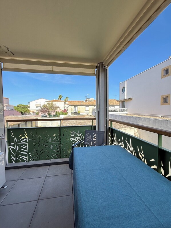 A balcony with a blue sofa, a green decorative fence, and a view of buildings and trees. - T2 avec terrasse, parking et cellier | La Roselière Le Grau-du-Roi (Le Grau-du-Roi)