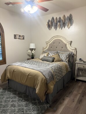 A bedroom with a bed, a nightstand, a lamp, a clock, a ceiling fan, and a wall decoration.
