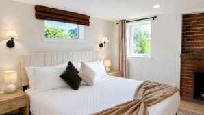 Ein ordentlich bezogenes Bett mit weißer Bettwäsche, einer braunen Decke und zwei Kissen. Zwei Nachttischlampen, ein Fenster mit Blick ins Grüne und ein Kamin aus Ziegelsteinen sind ebenfalls zu sehen.