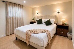 A bedroom with a bed, bedside lamps, a nightstand, a trash bin, and a window with curtains.