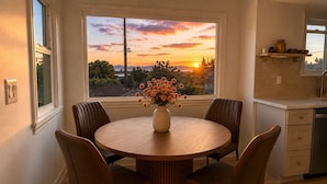 Ein Essbereich mit einem runden Tisch, vier Stühlen, einer Blumenvase und Blick auf einen Sonnenuntergang durch ein Fenster.