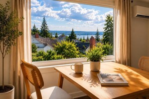 Quarto com vista para casas e árvores pela janela, uma mesa de madeira com uma planta e um livro, e uma cadeira.