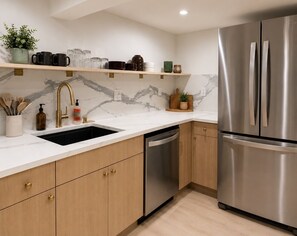 A modern kitchen with a stainless steel refrigerator, a sink with a gold faucet, wooden cabinets, and marble backsplash.