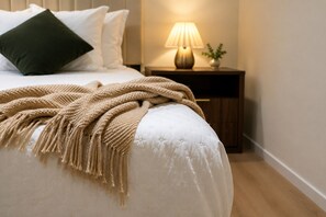 Cama bem arrumada com manta bege, luminária de cabeceira e uma pequena planta em mesa de cabeceira de madeira.