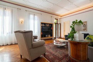 Een woonkamer met een beige fauteuil, een ronde salontafel, een boekenkast en een groot raam met witte gordijnen.