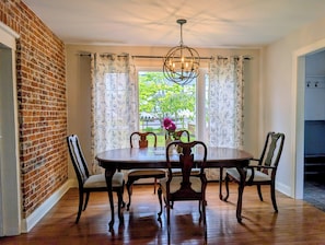 A dining room with a brick wall, a round table, four chairs, a chandelier, and a window with curtains.