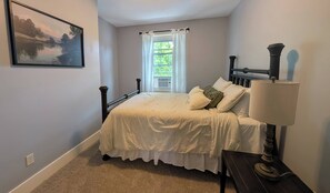 Quarto com cama, mesa de cabeceira, abajur, janela com cortinas e um quadro na parede.