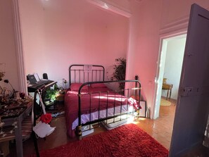 Een slaapkamer met een metalen bedframe, een rode sprei, een bureau met toetsenbord en een rood vloerkleed.