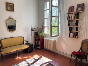 A room with a sofa, a chair, a radiator, a bookshelf, a window, and a mirror. - Character house with garden 10 minutes walk from the old town (aix en provence)