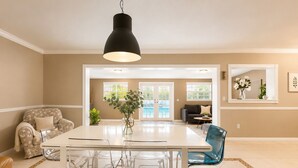 Uma área de refeições com uma mesa e cadeiras brancas, uma luminária pendente preta e vista para um espaço ao ar livre através de uma porta de vidro.