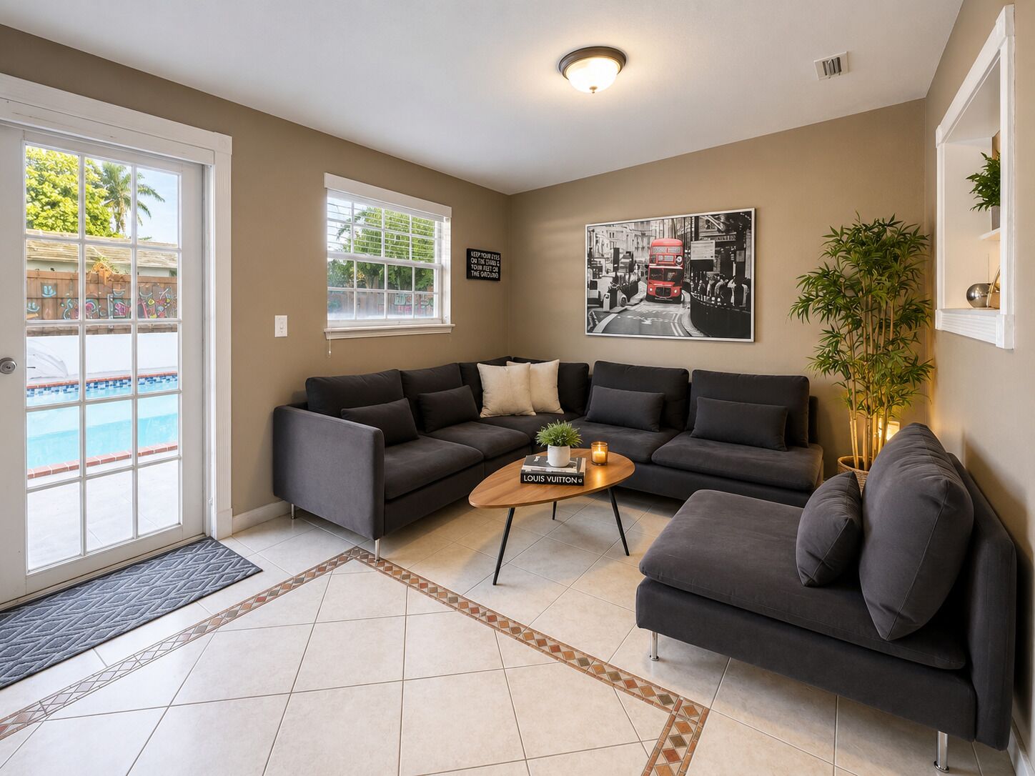 Sala de estar con sofá gris, una mesa de centro, una planta en maceta y vista a la piscina a través de una puerta de vidrio.