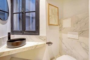 A bathroom with a marble countertop, a round sink, a window with a black frame, a framed abstract artwork, and a wall-mounted light switch. - ELEGANT 3BR APARTMENT WITH ENSUITE BATHS NEAR CANAL SAINT-MARTIN (Paris)