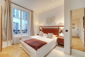 A bedroom with a large bed, a window with curtains, a radiator, and a wooden floor. - ELEGANT 3BR APARTMENT WITH ENSUITE BATHS NEAR CANAL SAINT-MARTIN (Paris)