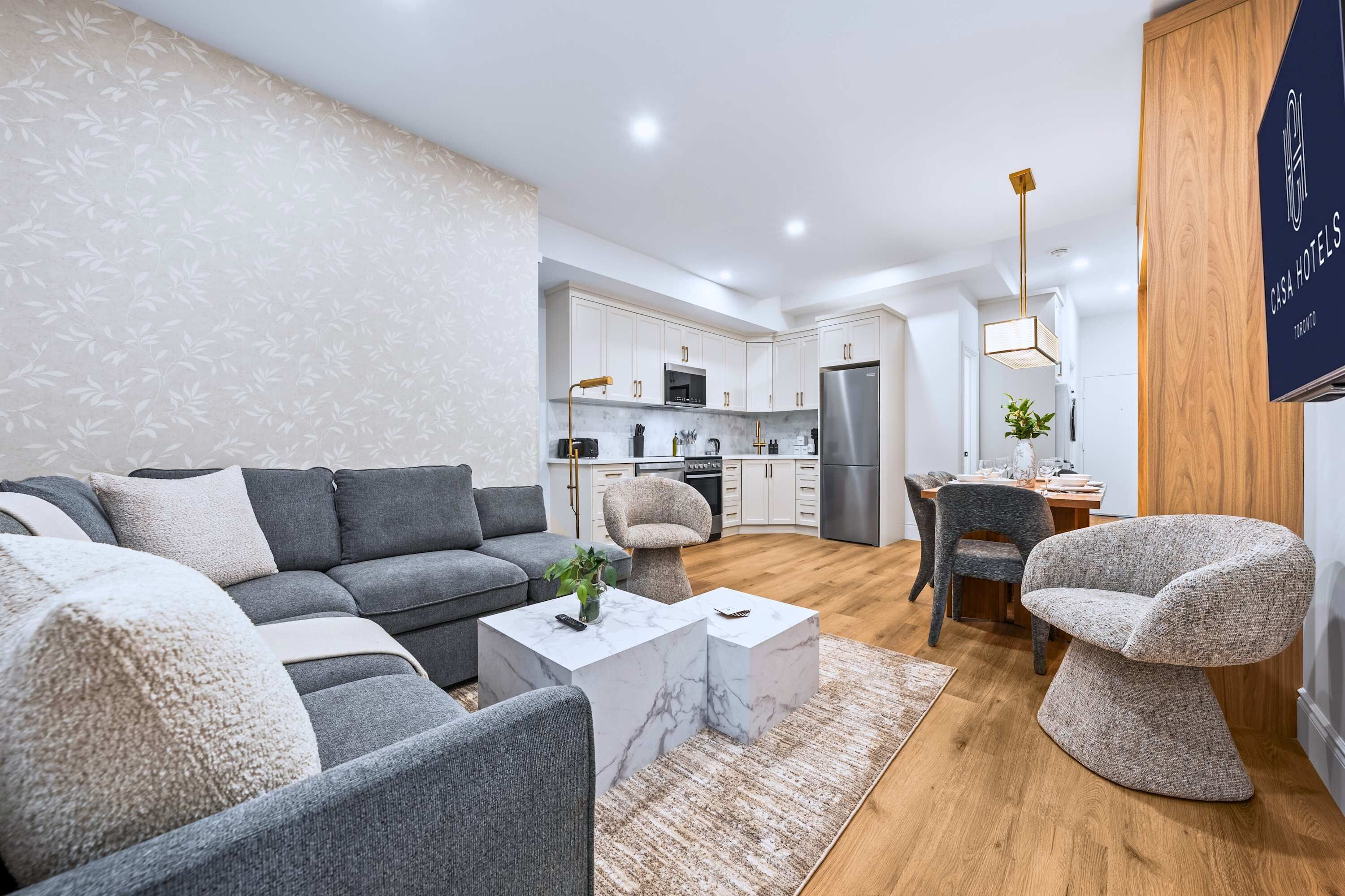 Una sala moderna con un sofá modular gris, una mesa de centro de mármol y una silla original. Se ve una cocina con armarios blancos y electrodomésticos de acero inoxidable al fondo.