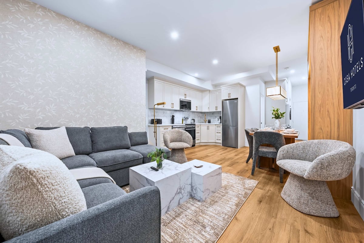 Una sala moderna con un sofá modular gris, una mesa de centro de mármol y una silla original. Se ve una cocina con armarios blancos y electrodomésticos de acero inoxidable al fondo.