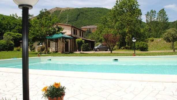 A swimming pool with a tiled deck, a lamp post, and a building with a green roof in the background. - Il Bosso Apartment in Cagli with a 15x6m saltwater pool (Cagli)