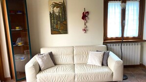A beige sofa with cushions, a painting on the wall, a radiator, and a window with curtains. - Il Bosso Apartment in Cagli with a 15x6m saltwater pool (Cagli)