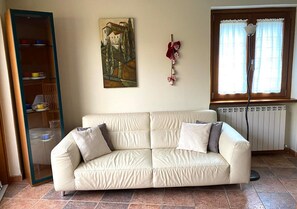 Een beige bank met kussens, een schilderij aan de muur, een radiator en een raam met gordijnen.