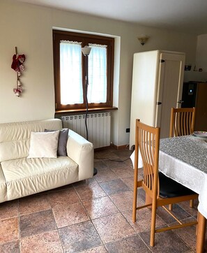 Een kamer met een witte bank, een eettafel met stoelen, een radiator, een raam met gordijnen en een wandlamp.