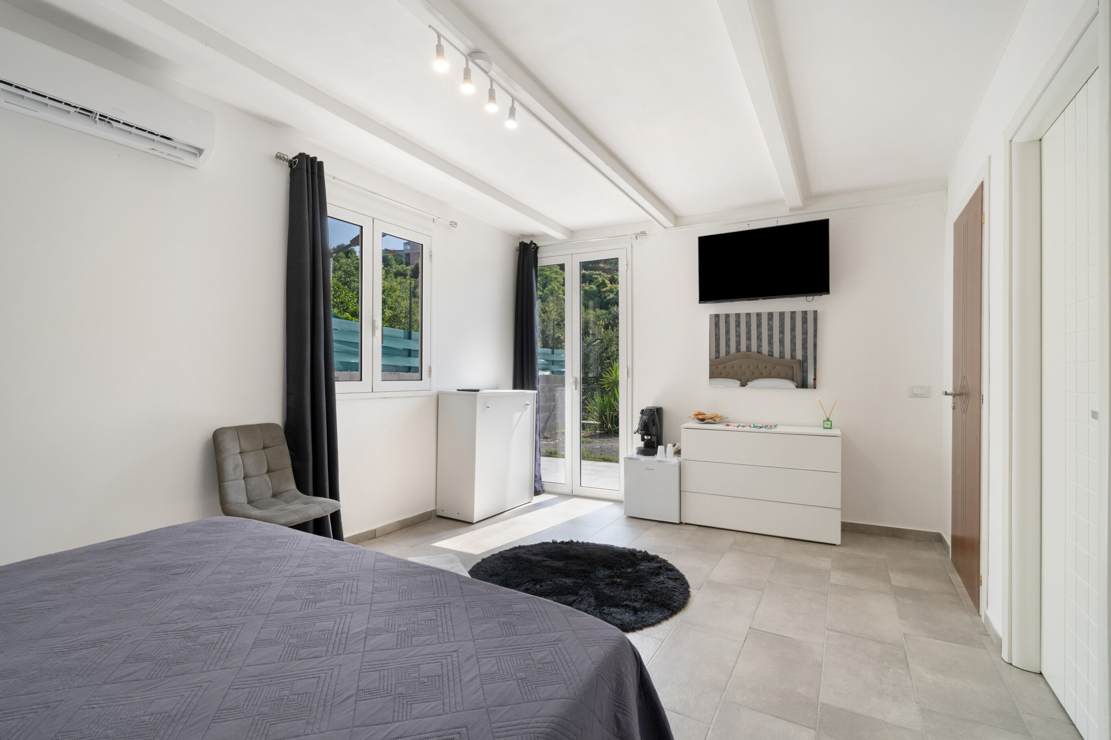 A modern bedroom with a bed, a desk, a television, and a view of greenery outside.