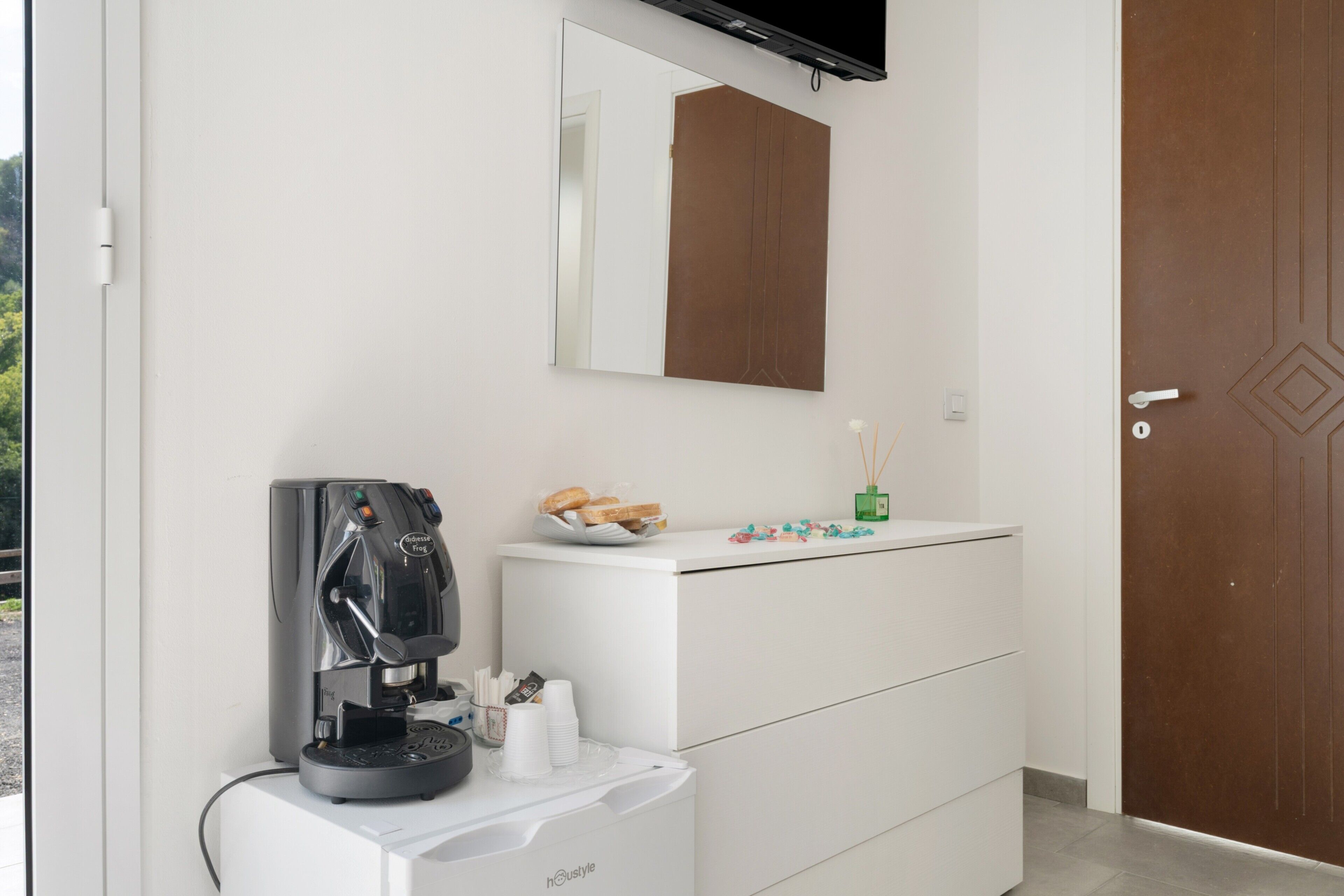 A modern bathroom with a coffee machine, a mirror, and a white cabinet with a tray of bread and condiments.