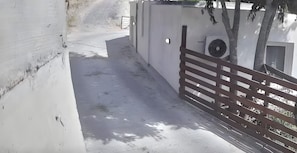 A narrow pathway leading to a building with a white exterior, a metal gate, and a ventilation unit on the wall.