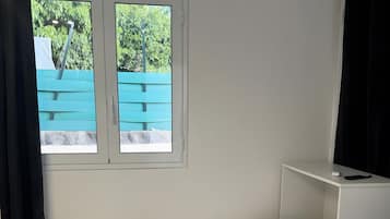 A single bed with white bedding, a small bedside table, a window with a view of greenery, and a curtain.