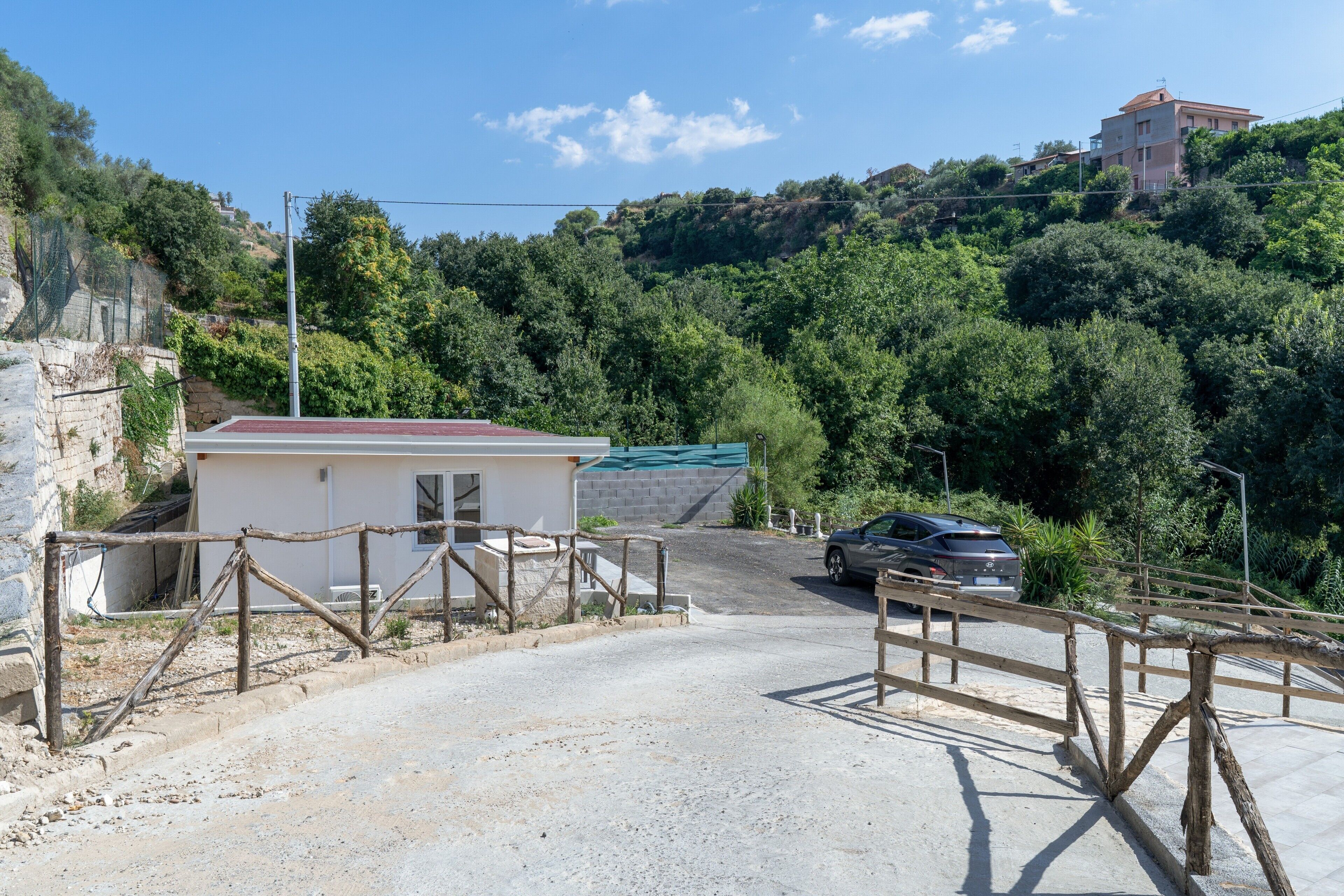 A gravel driveway leading to a building with a red roof, surrounded by greenery and a hill in the background.