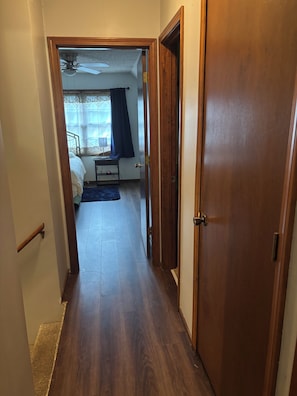 Un couloir étroit mène à une chambre qui contient un lit, une table de chevet et un ventilateur de plafond.