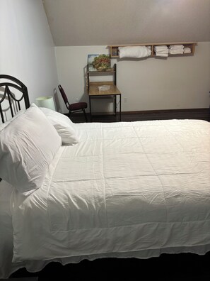 Ein ordentlich bezogenes Bett, ein Holztisch mit Stuhl und ein Regal mit gefalteten Handtüchern.