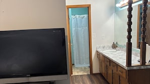 Una habitación con televisión, armarios de madera, un baño con cortina de ducha y un espejo con luces.