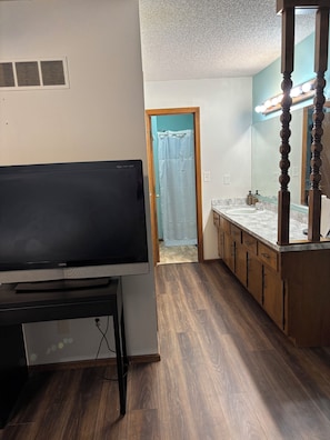 Une chambre dotée d’une télévision, de meubles en bois, d’une salle de bain avec un rideau de douche et d’un miroir avec éclairage.