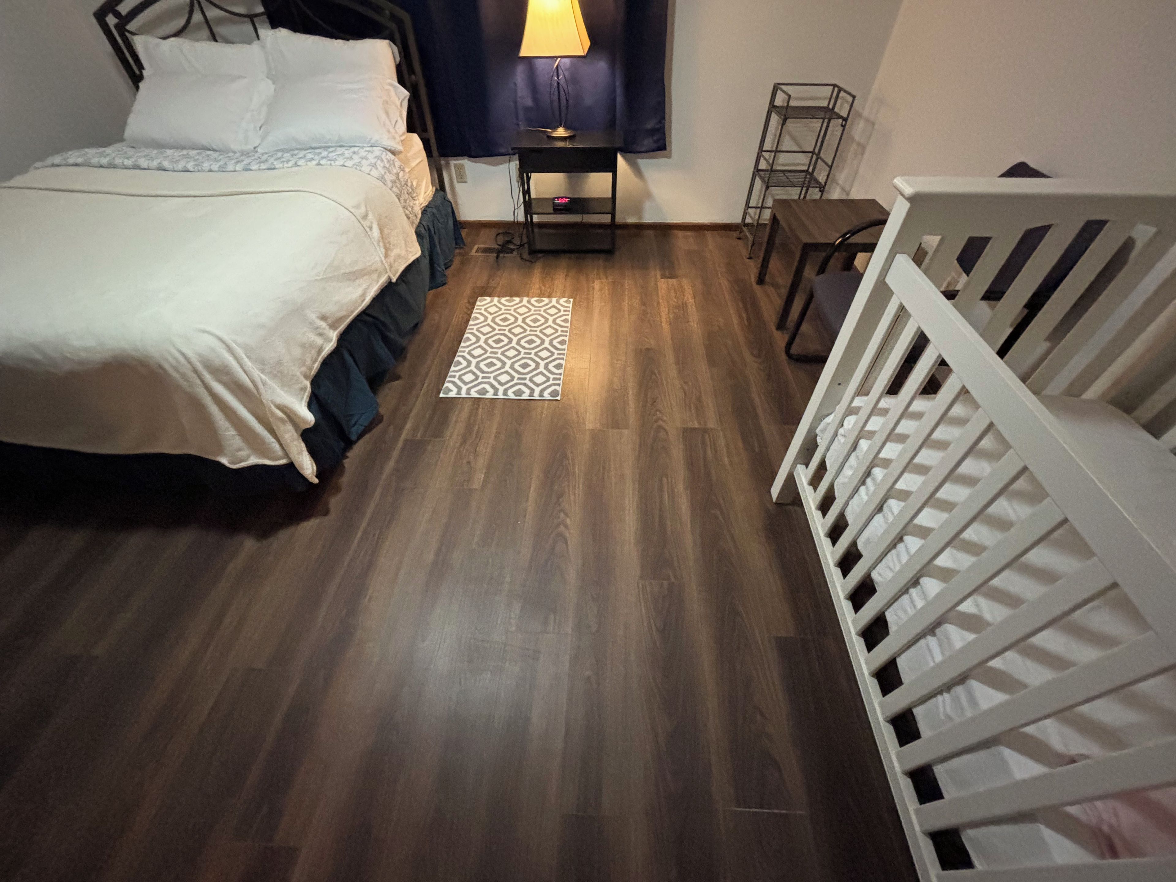 A bedroom with a bed, a nightstand, a lamp, a crib, and a patterned rug.