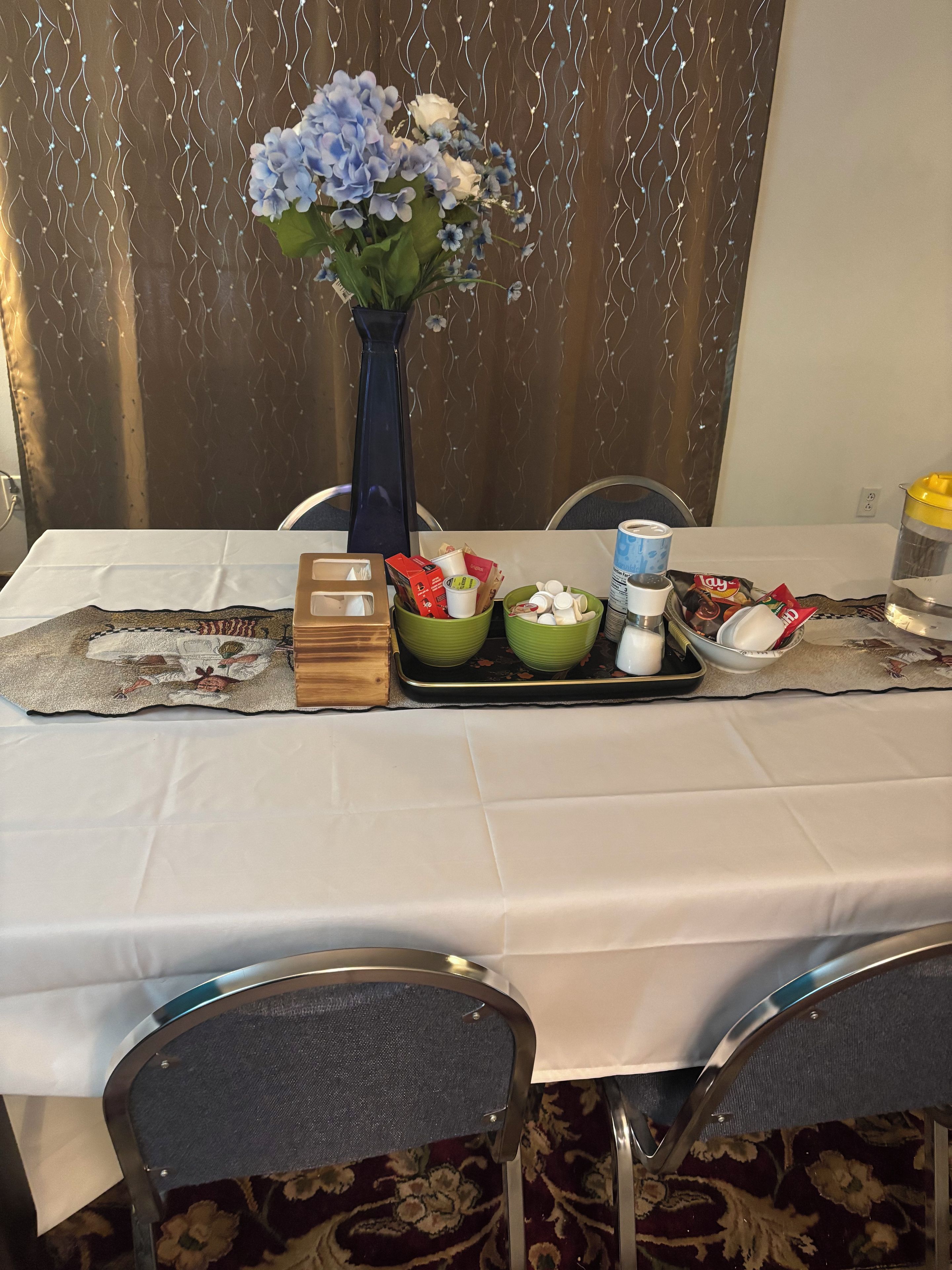 A table set with a vase of flowers, various food items, and a tray with condiments.
