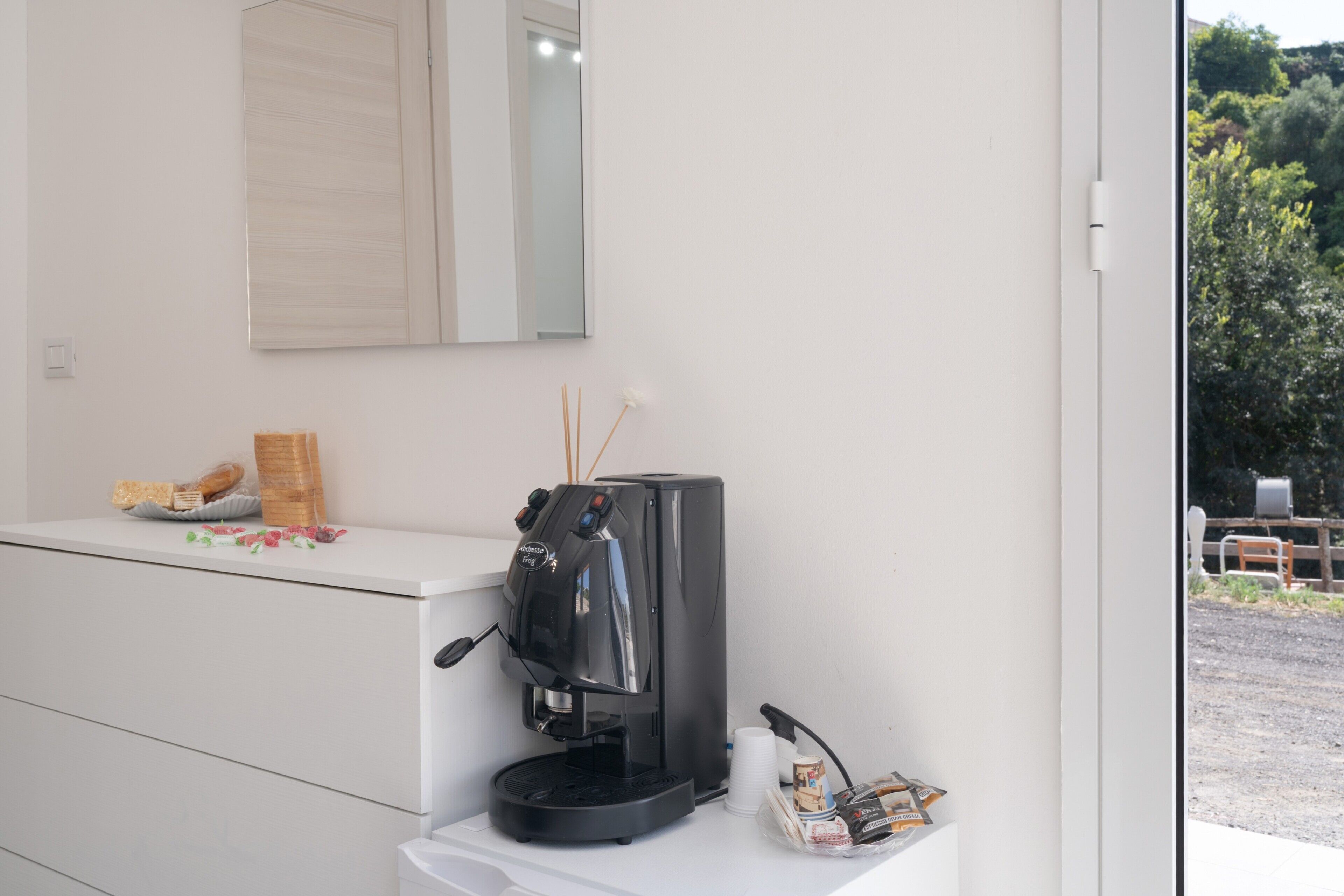 A coffee machine on a white counter with a mirror above it, a window with a view of trees, and a door to the right.