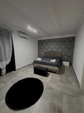 A bedroom with a bed, a black ottoman, a black furry rug, and a patterned wallpaper.
