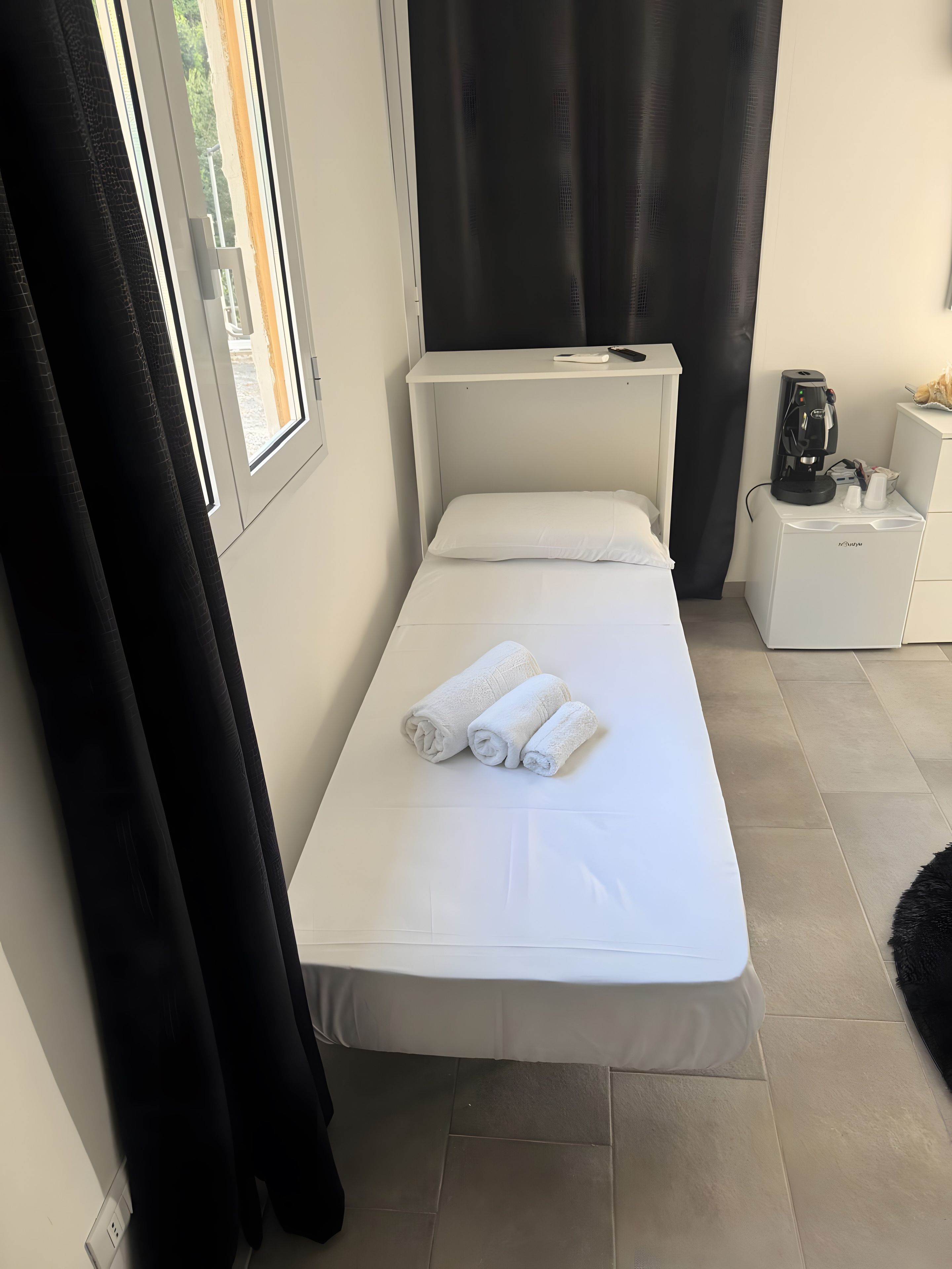 Una cama individual con sábanas blancas y toallas enrolladas, una mesita de noche con cafetera y una ventana con cortinas.