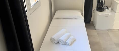A single bed with white linens and rolled towels, a bedside table with a coffee maker, and a window with curtains.