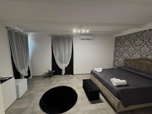 A bedroom with a bed, a small bedside table, a black rug, a potted plant, and a window with sheer curtains.