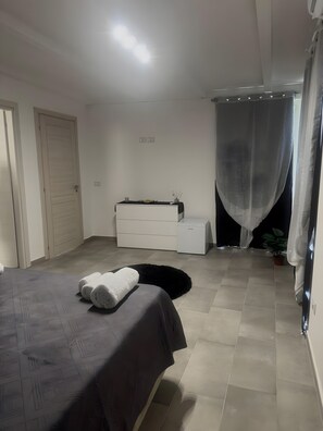 A bedroom with a bed, a dresser, a small table, a potted plant, and a window with sheer curtains.