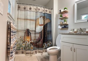 Ein Badezimmer mit Duschvorhang, der einen Cowboyhut und Blumen zeigt, einer Toilette und einem Schminktisch mit Spiegel und Ablageflächen.