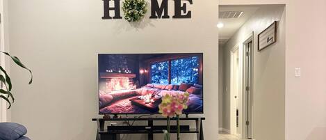 A living room with a flat-screen TV, a coffee table with a book and a plant, a blue sofa, and a 'HOME' sign on the wall.